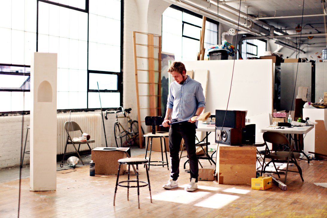A male student in a blue shirt works on a design project in a bright, industrial sculpture studio filled with materials and tools.