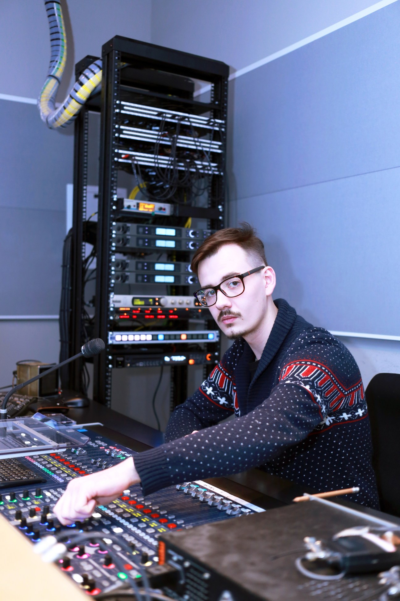 Sound director Peter Suskov sits at a control desk with audio equipment, facing a server rack with networking cables above.