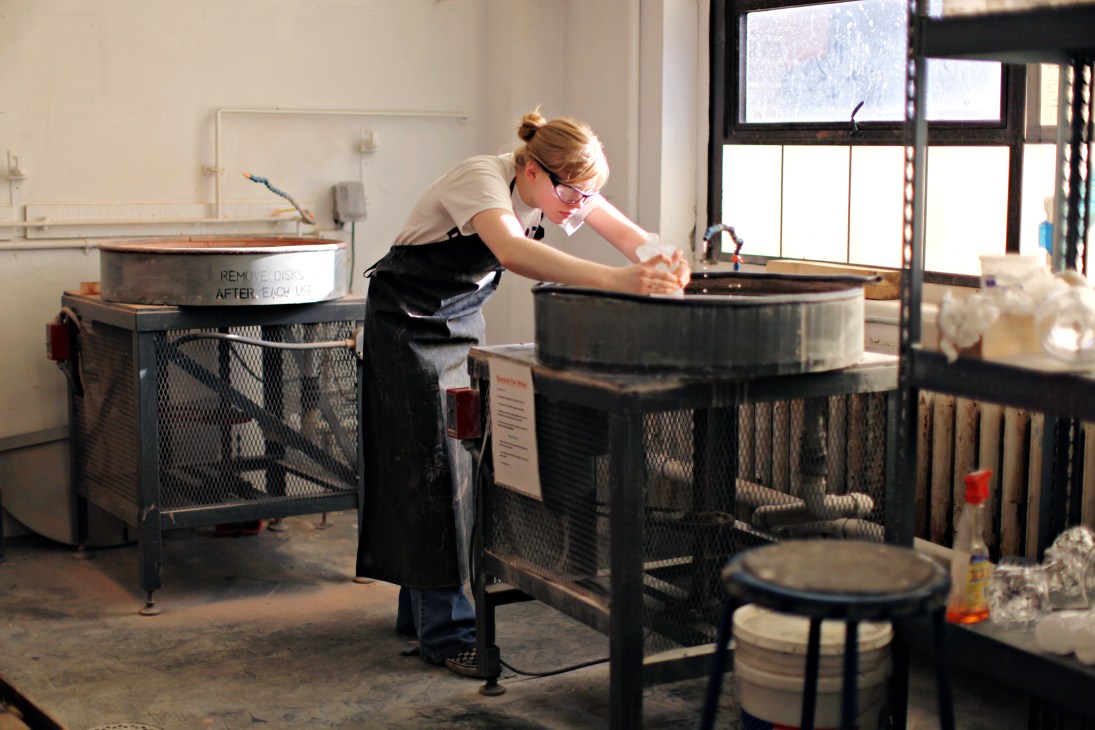 Student Hannah Kirkpatrick works with molten glass in a studio hot shop surrounded by equipment.