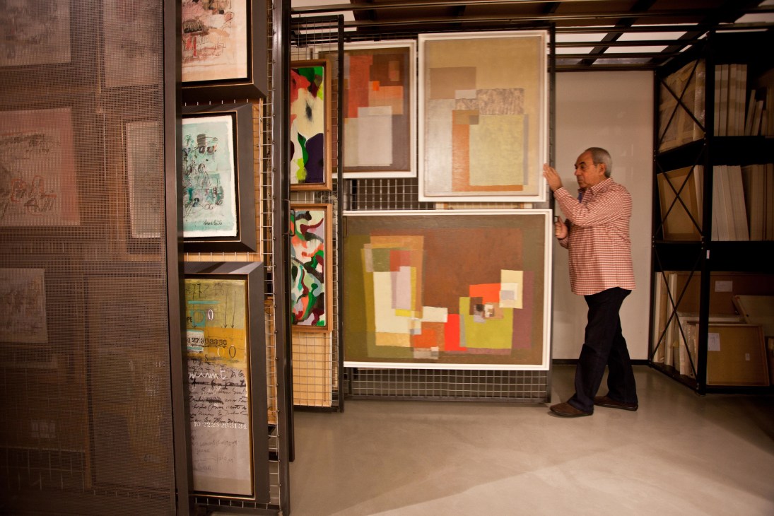 A man in a pink striped shirt examines framed abstract artworks displayed in a storage room with industrial shelving.