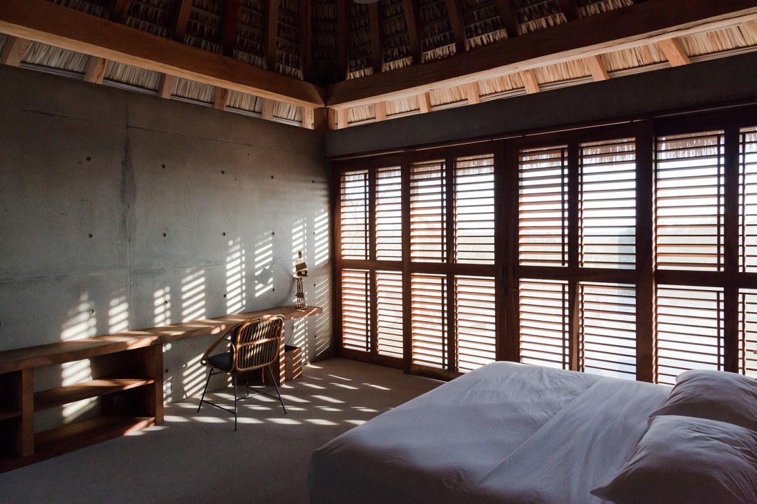 Minimalist beach cabana interior with concrete walls, wooden shutters, desk, chair and bed with natural light.