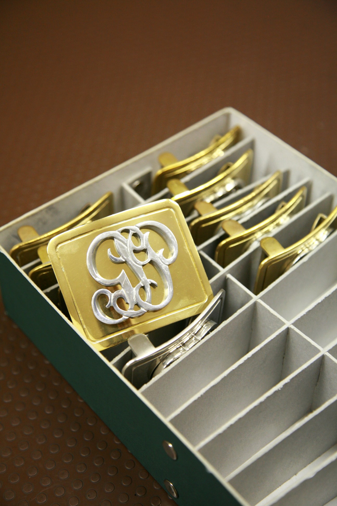 Brass belt buckles with ornate silver insignia stored in a compartmentalized white box.