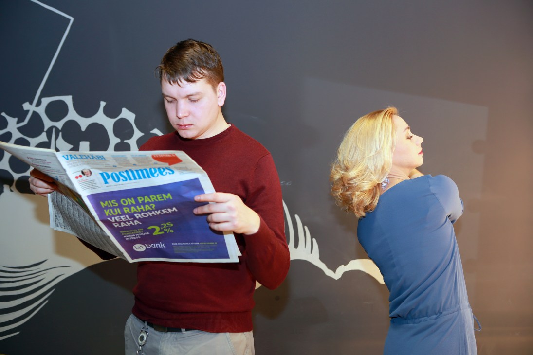 A boy in a maroon sweater reads a Russian-language newspaper while a woman in a blue dress stands beside him.
