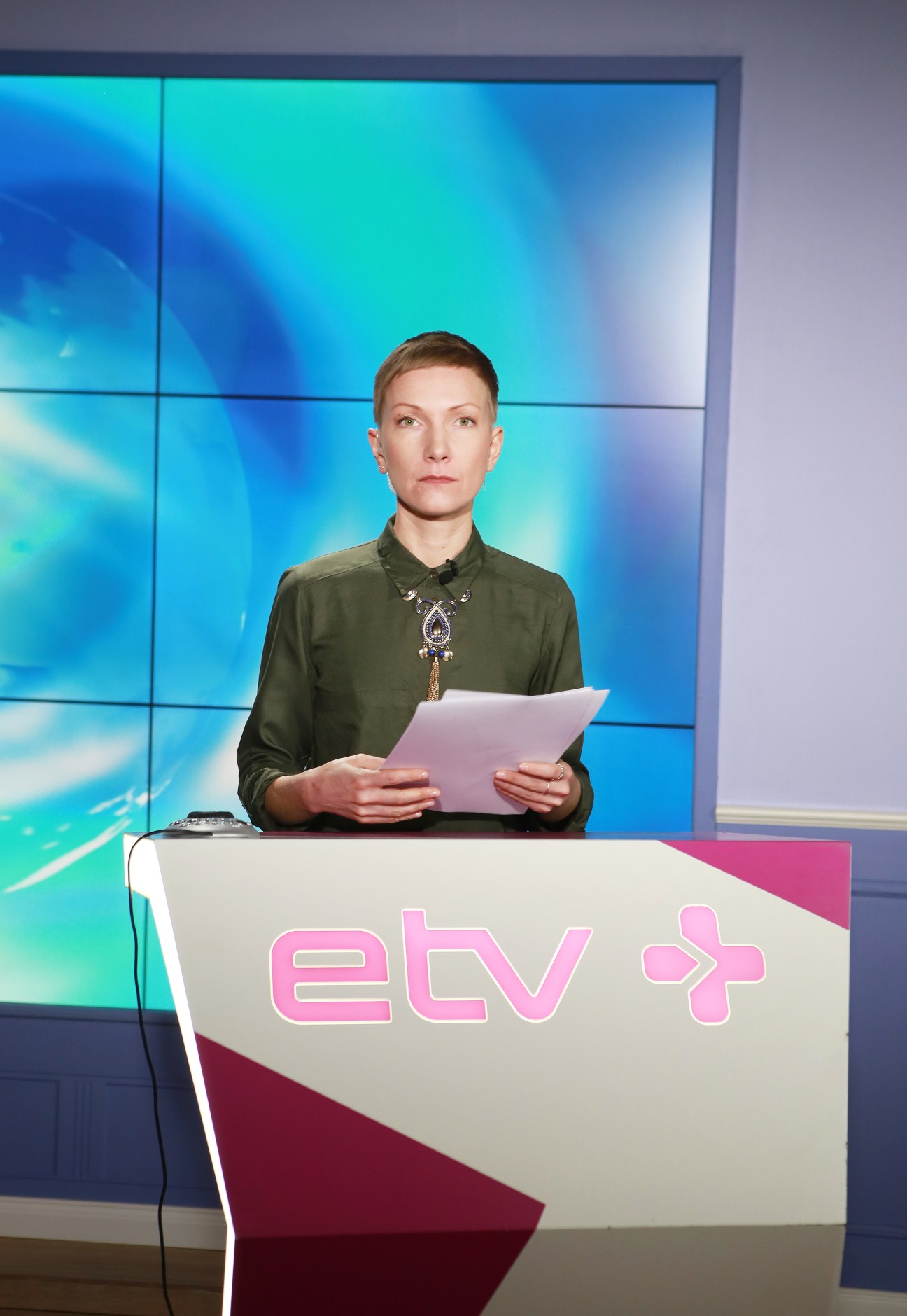 Newsreader Tatiana Kosmynina at ETV+ desk holding script with blue video wall background.