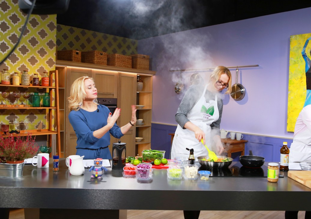 Two women cooking together on a television set with colorful kitchen backdrop and fresh ingredients on counter.
