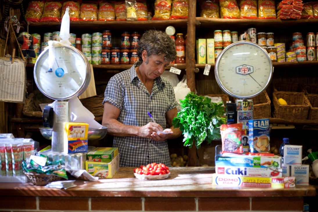 Shopkeeper at I Fruttarelli fruit and vegetable store in Genoa arranging fresh produce at the counter.