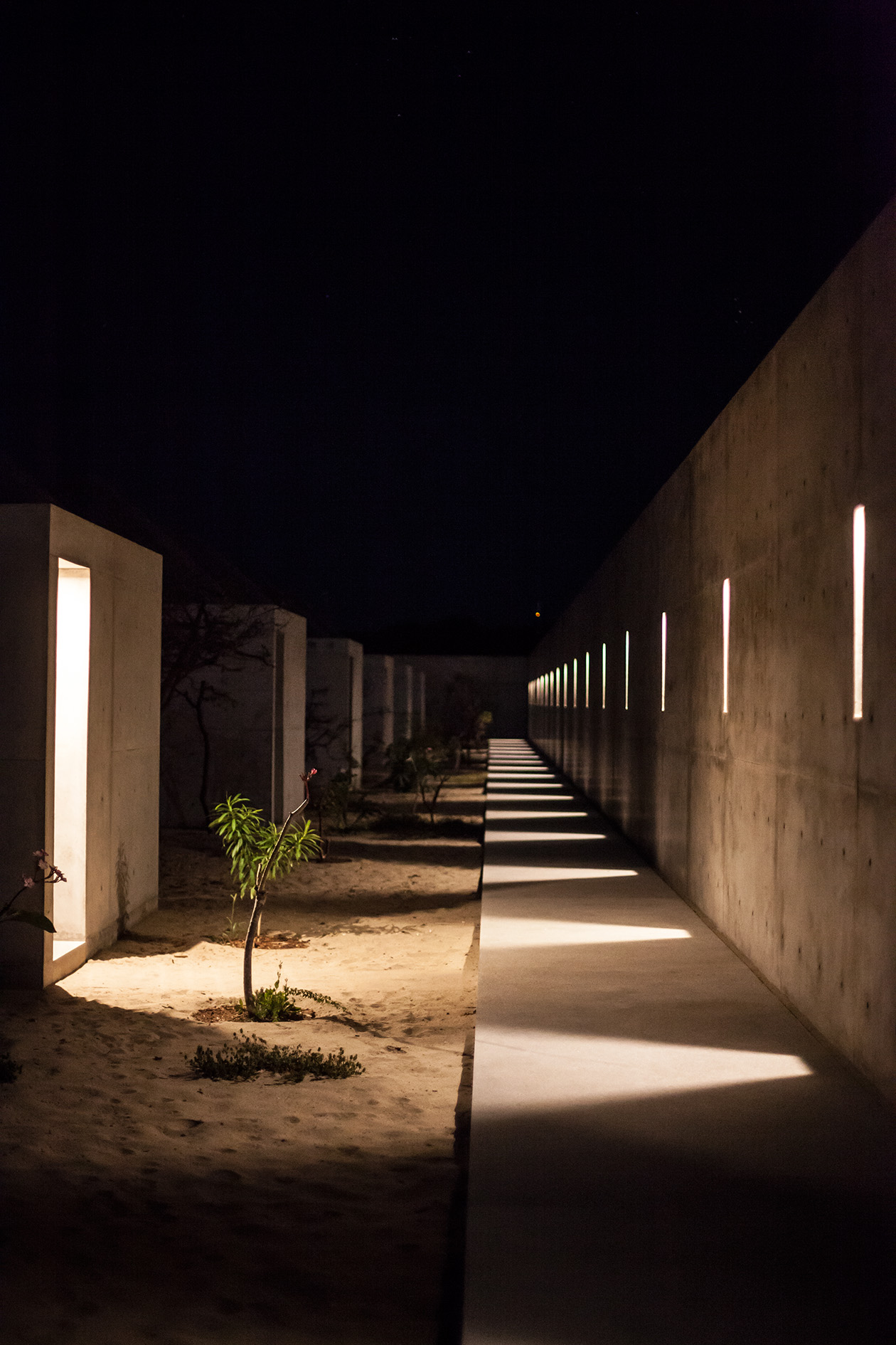 Illuminated concrete cabanas line a sandy path at night, with geometric light fixtures creating dramatic shadows.