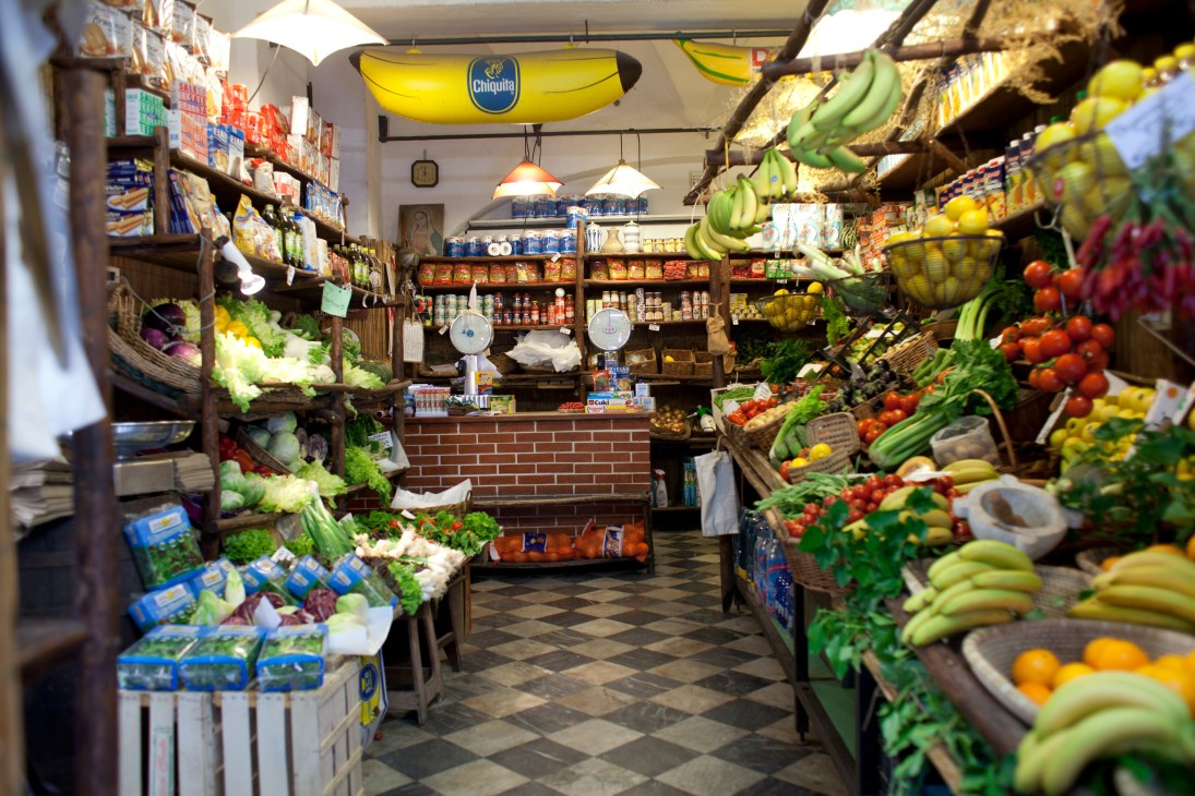 Interior of I Fruttarelli fruit and vegetable shop in Genoa displaying fresh produce on wooden shelves and hanging bananas.
