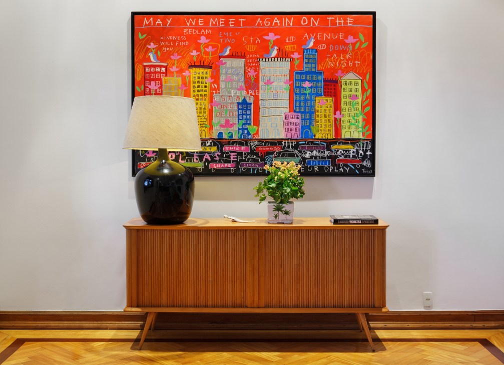 Colorful artwork depicting city buildings and street scenes hung above a mid-century wooden sideboard with a black lamp and potted plant.