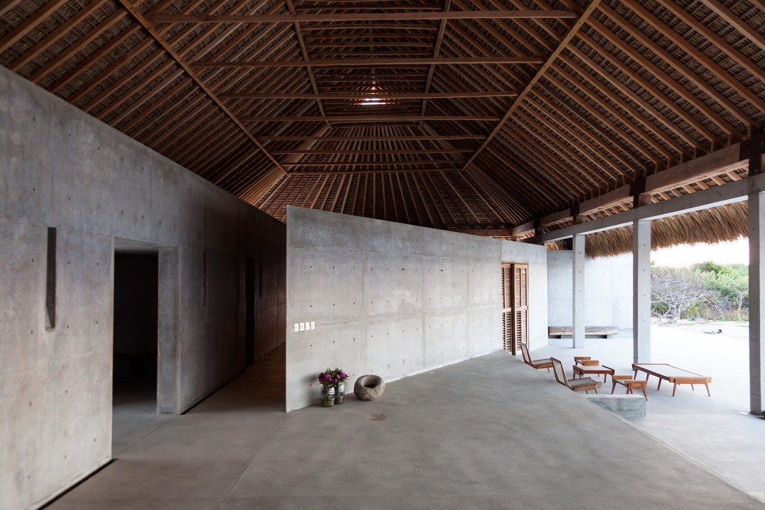 Interior of concrete main villa with thatched-palm roof, open doorways, wooden chairs, and ocean view.