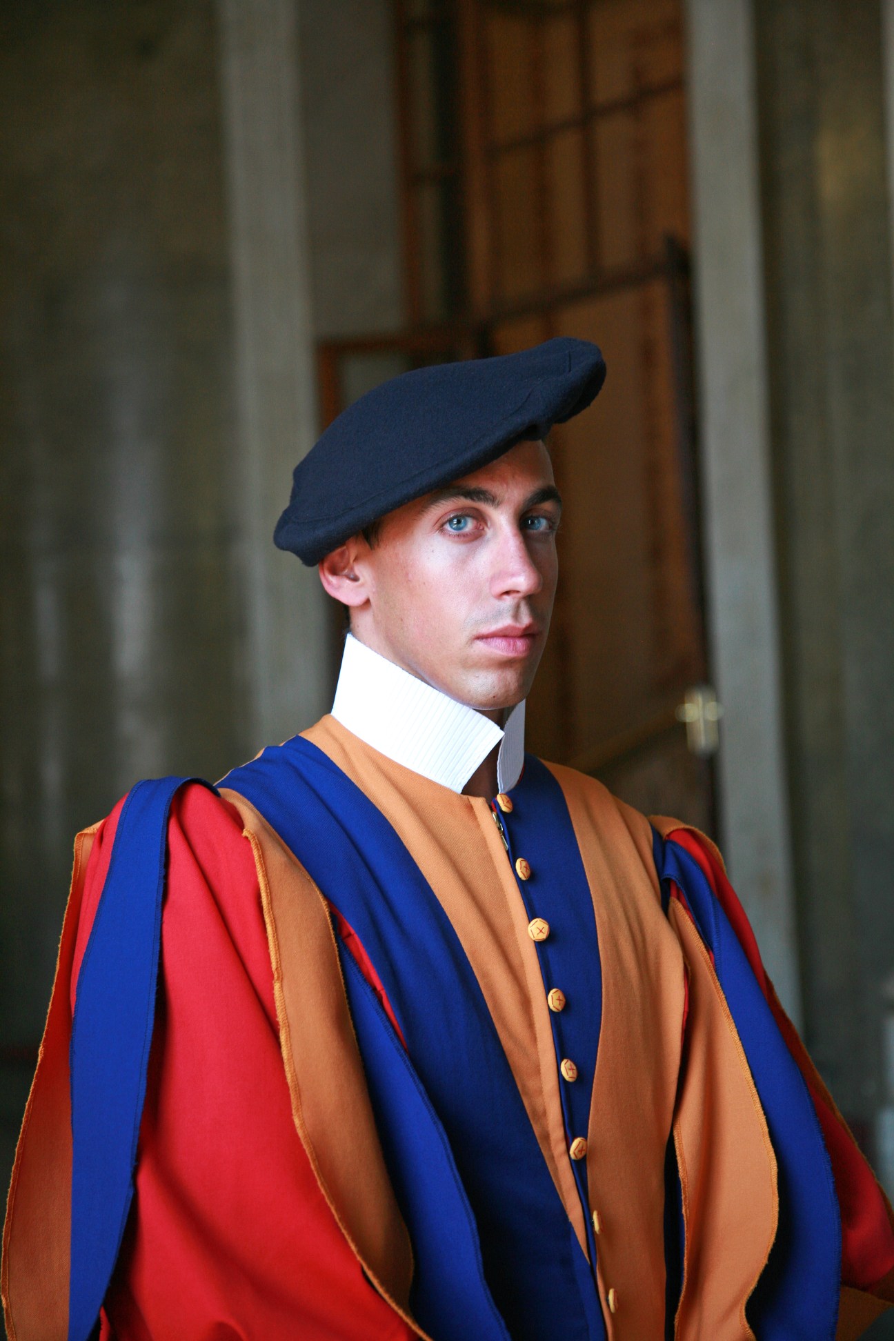 Guardsman Marco Ruppen in formal Swiss Guard dress uniform with blue and yellow striped jacket, red cape, white collar, and pleated beret.
