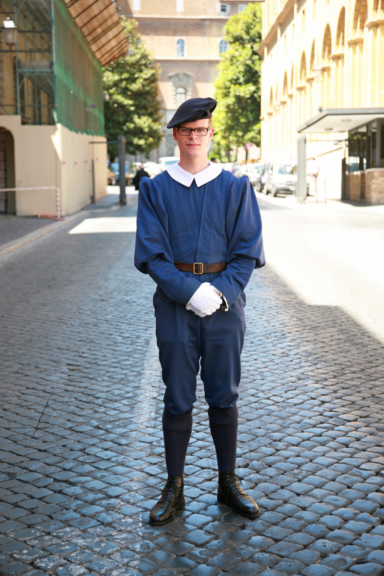 Swiss Guard in blue night uniform with white collar and beret standing on Vatican cobblestone street.