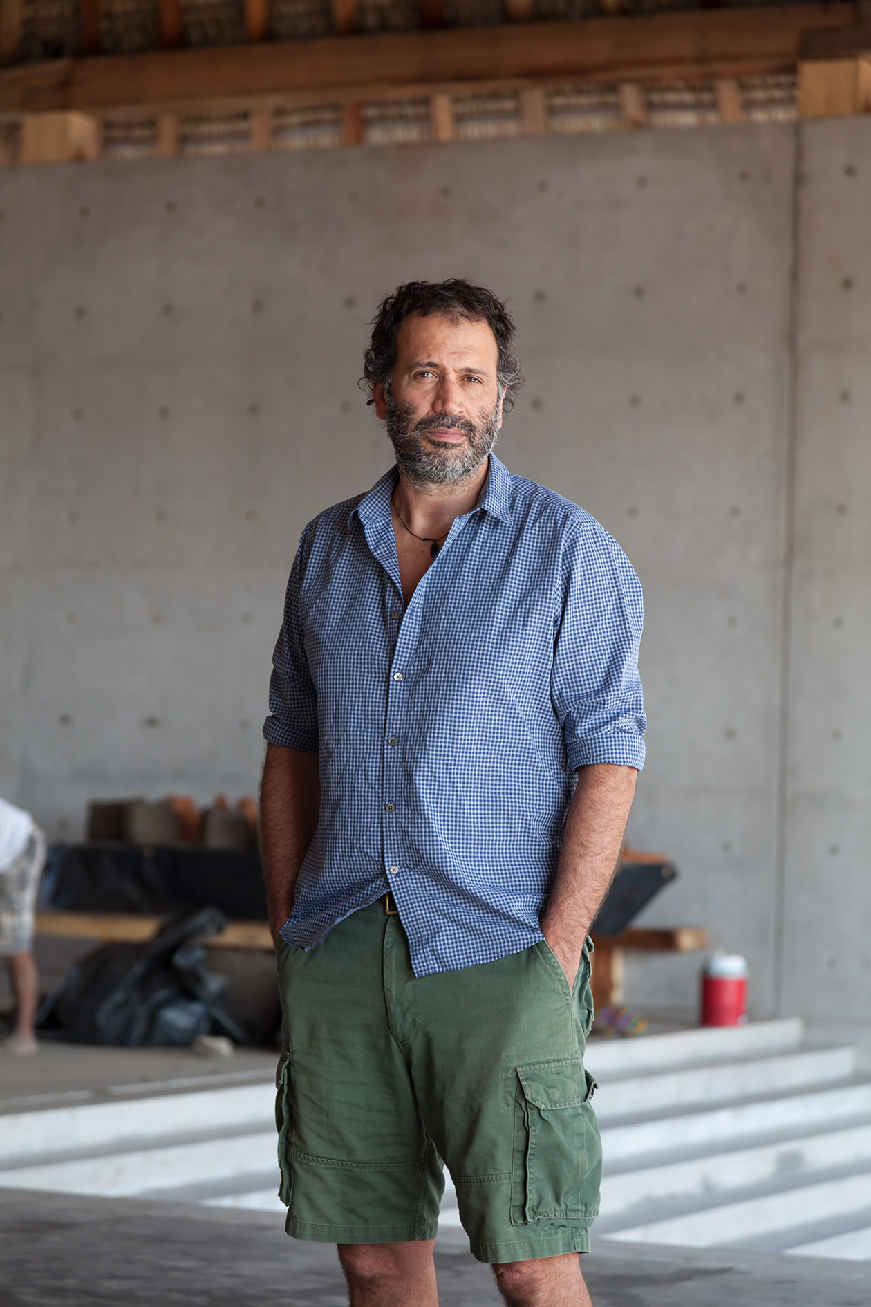 Mexican artist Bosco Sodi stands in a concrete studio space wearing a blue checkered shirt and green cargo shorts.