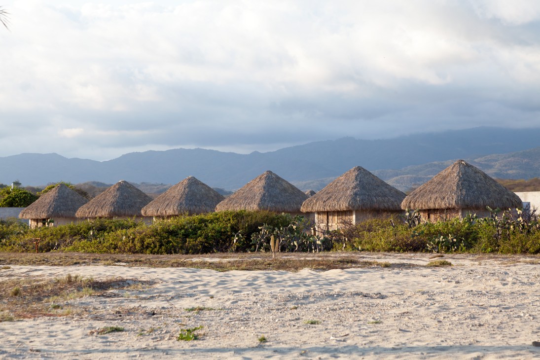 A row of five thatched-roof cabanas with cone-shaped roofs line a sandy beach with mountains in the background.