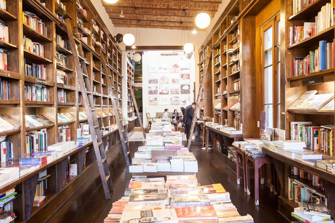 Interior of Libros del Pasaje bookshop with floor-to-ceiling wooden shelves stocked with books, library ladder, and customers browsing in the background.