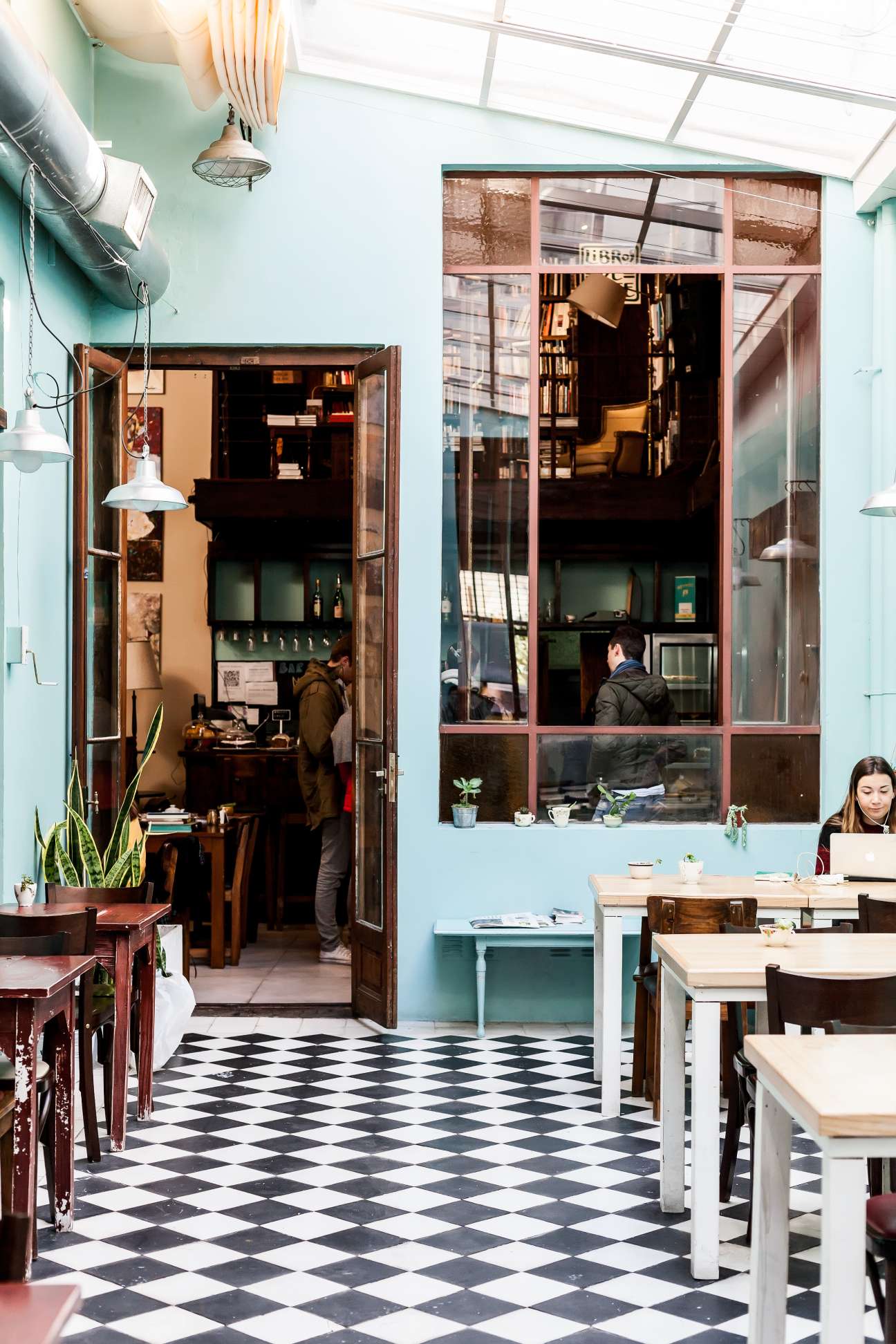 Light-filled café space with black and white diamond floor, mint green walls, and customers seated at wooden tables near a large window.