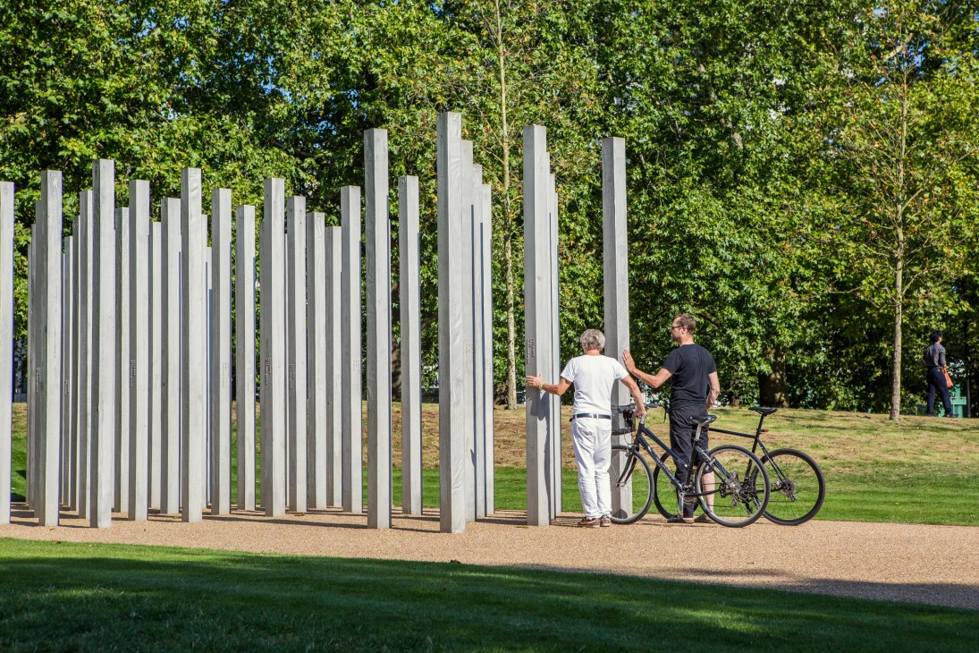 Two visitors interact with tall stainless steel stelae in Hyde Park, London, with trees in the background.