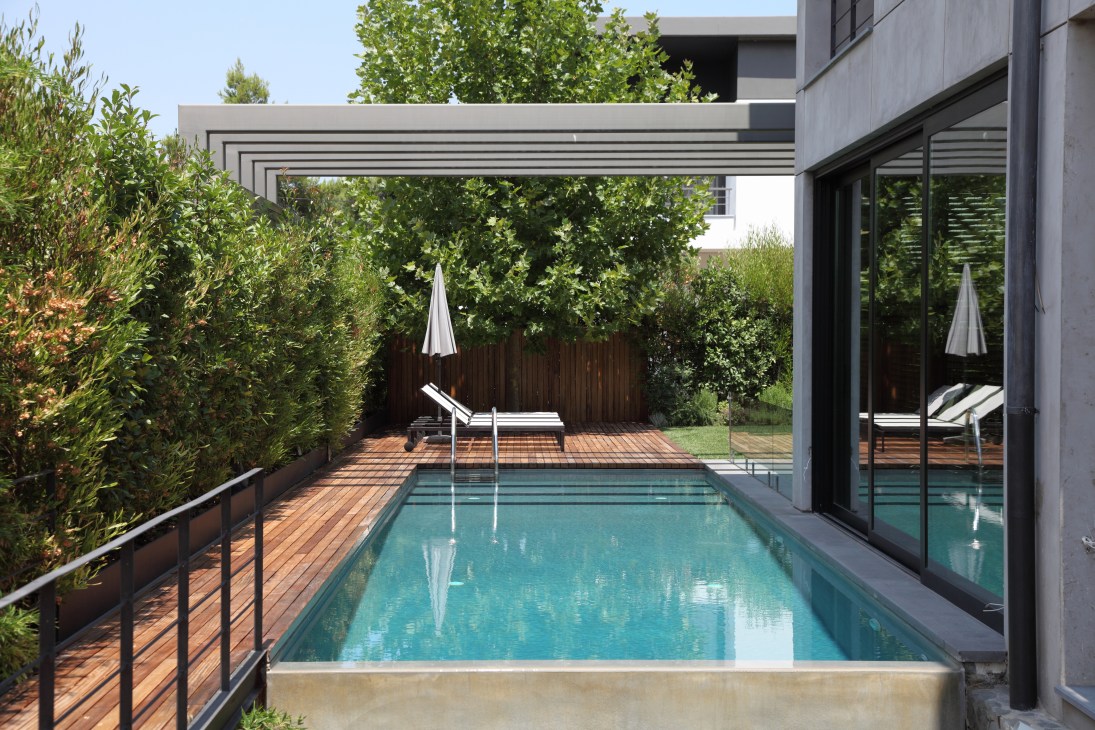 Modern residential pool surrounded by iroko wood decking with pergola and green landscaping.