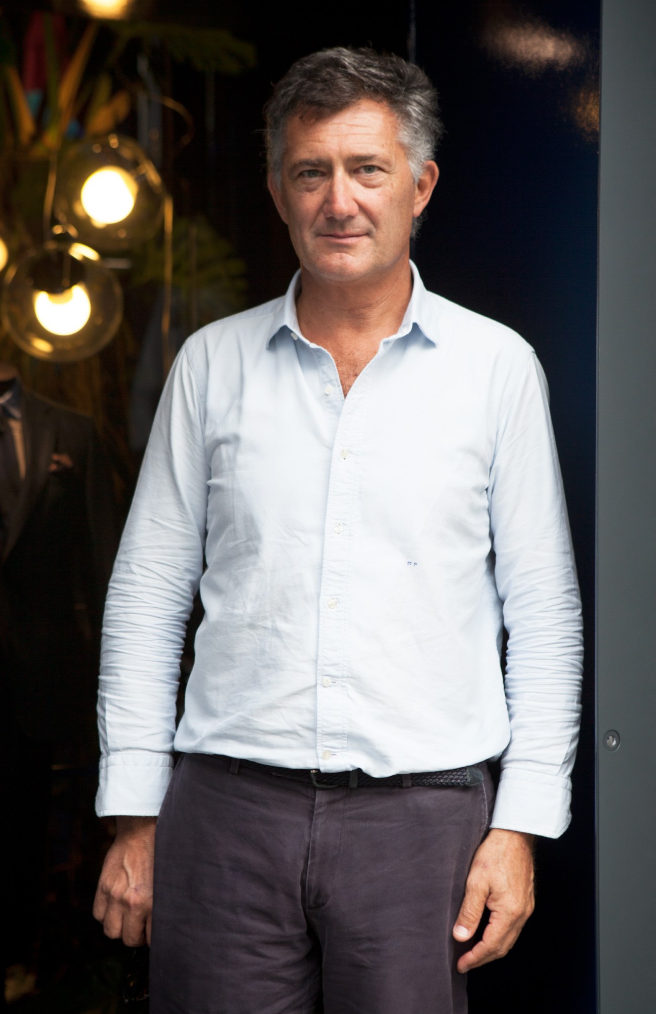Massimo Piombo wearing a light blue button-up shirt and dark trousers, standing in front of brass lighting fixtures.