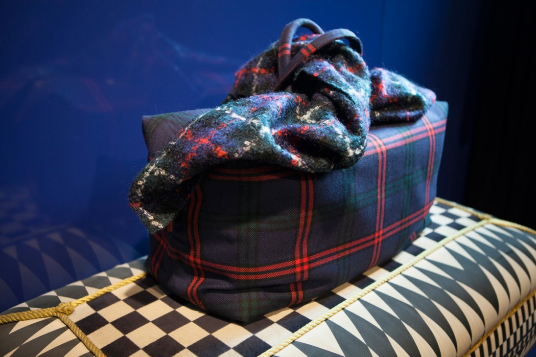 Massimo Piombo tartan wool garment draped over patterned decorative objects against blue background.