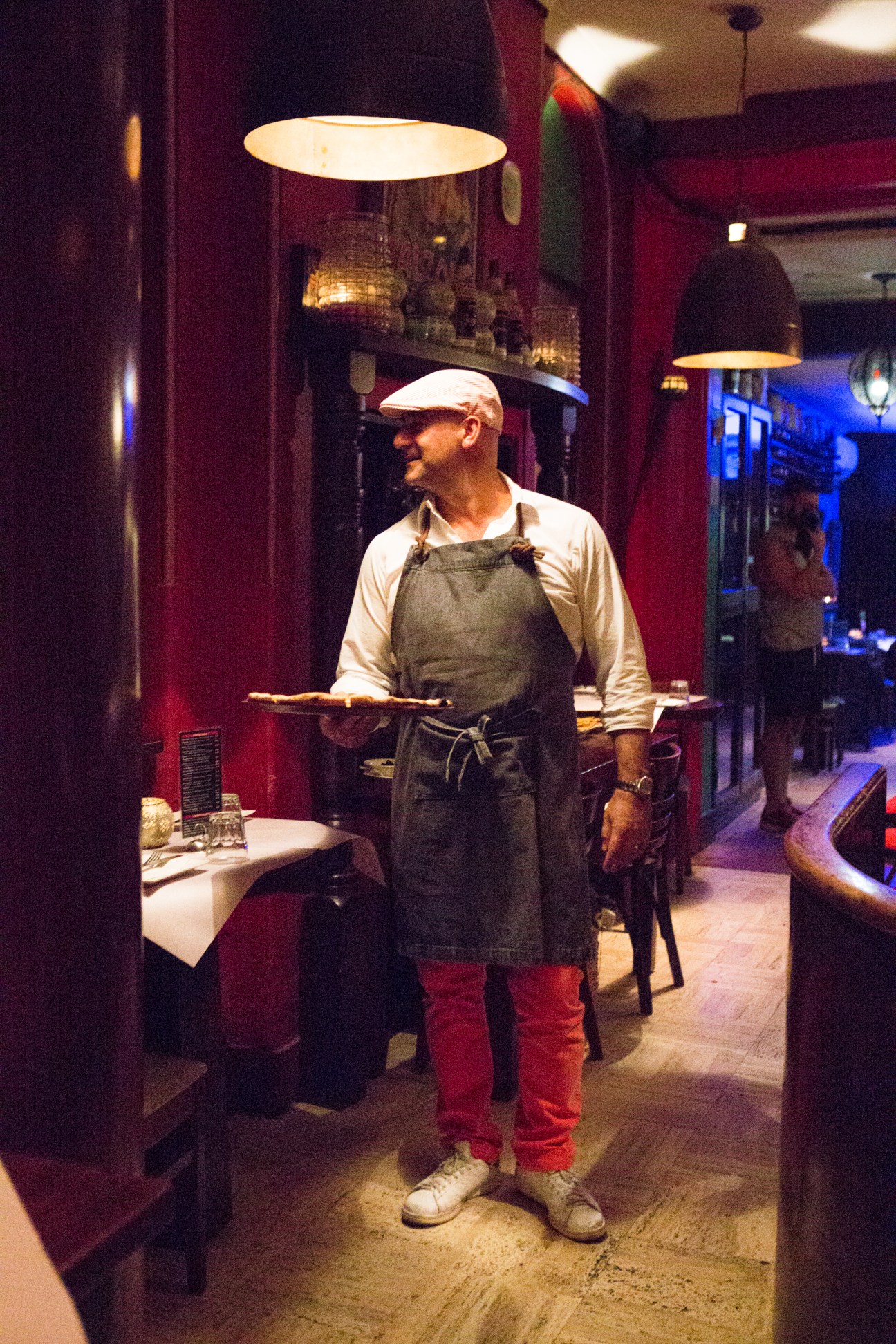 A server in an apron and cap holds a plate in a dimly lit restaurant with burgundy walls and pendant lighting.