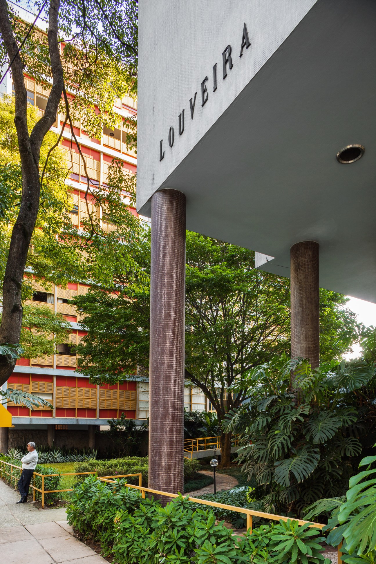 Exterior of Edificío Louveira showing modernist architecture with brick columns, signage, and surrounding tropical vegetation.