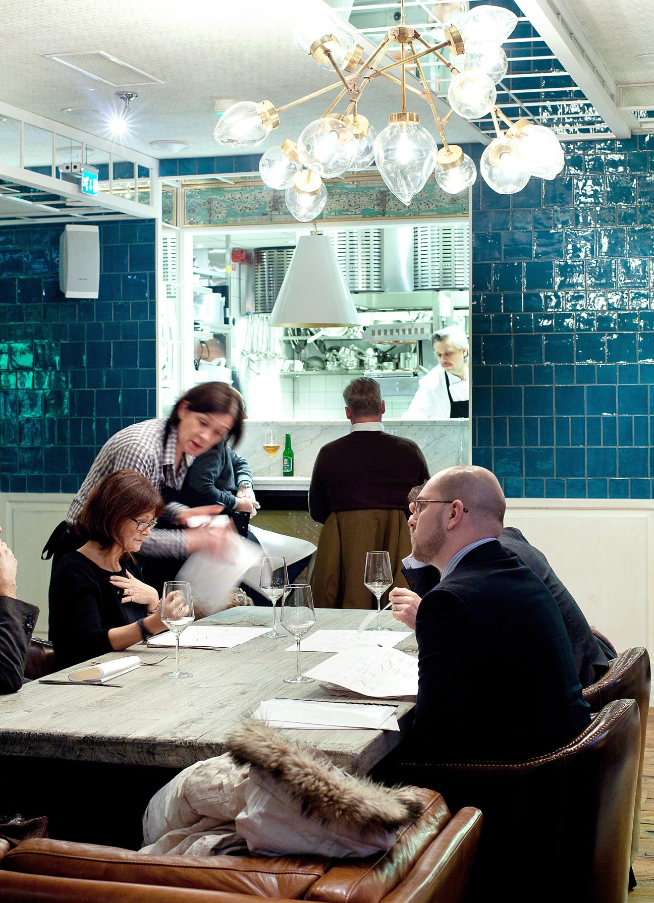 Restaurant interior at Nosh and Chow with diners seated at marble table and server in motion.