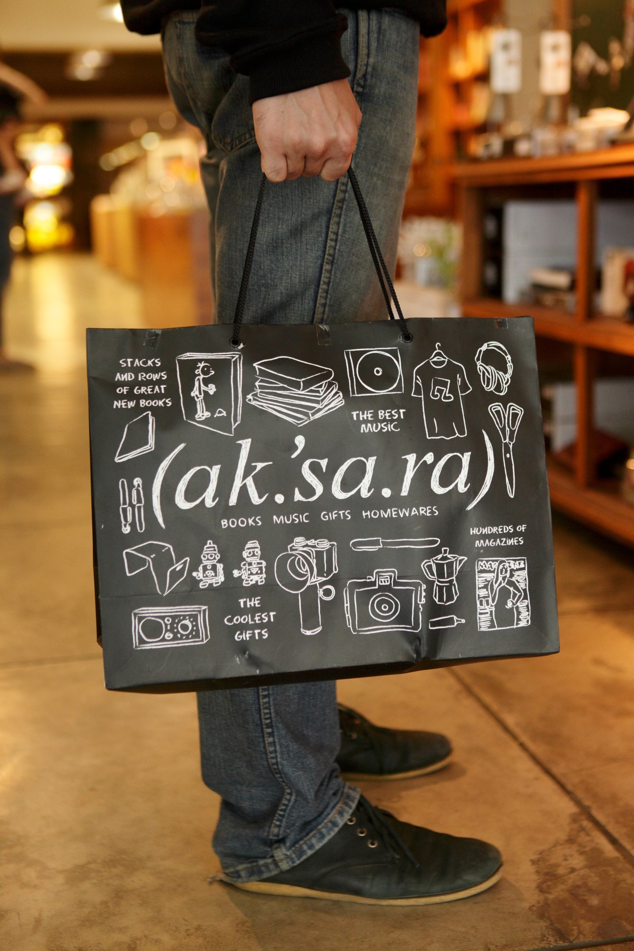 Person holding black Aksara shopping bag with white hand-drawn illustrations and text inside bookstore.