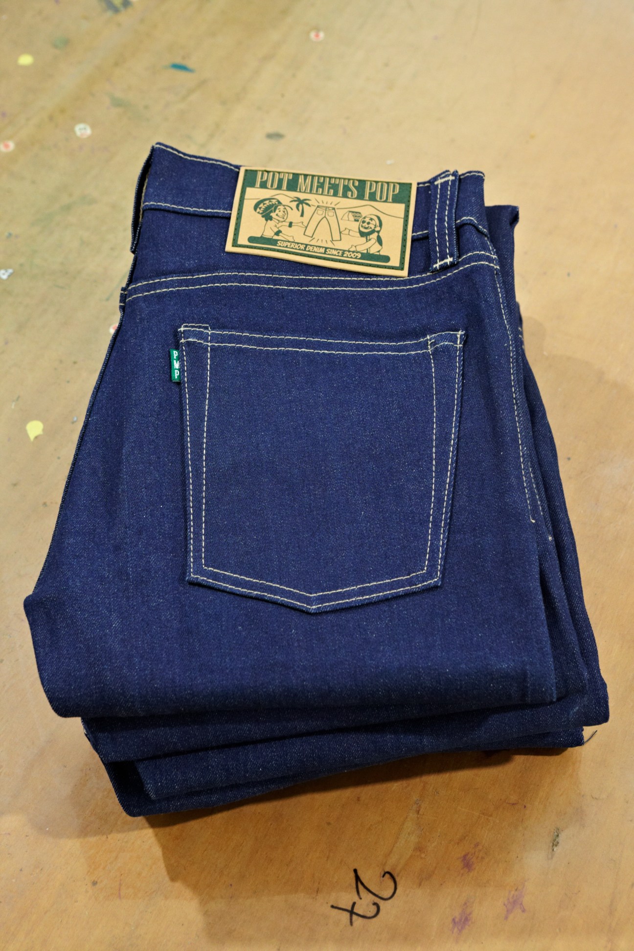 Folded dark indigo jeans with white stitching and Pot Meets Pop label displayed on tan surface.