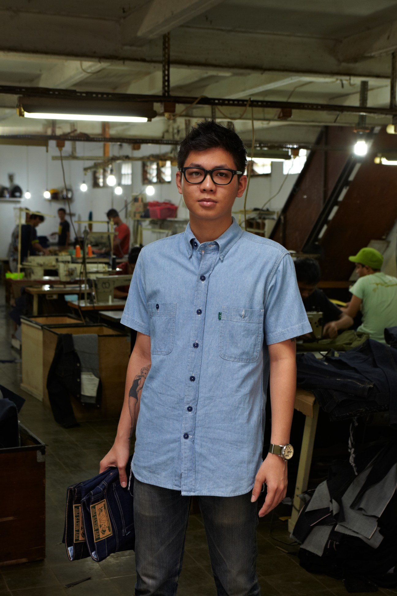 Hendry Sasmitapura stands in a denim workshop wearing a light blue shirt and holding fabric samples.