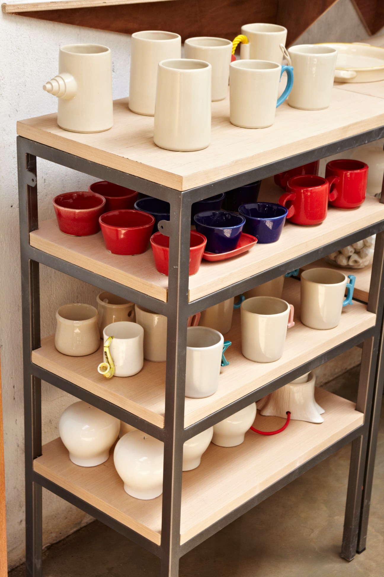 Shelving unit displaying handmade ceramic mugs and bowls in cream, red, and navy colors with decorative handles.