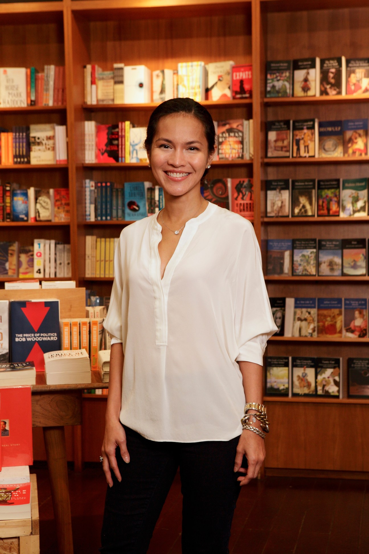 Woman in white shirt stands smiling in front of wooden bookshelves at Aksara bookshop.