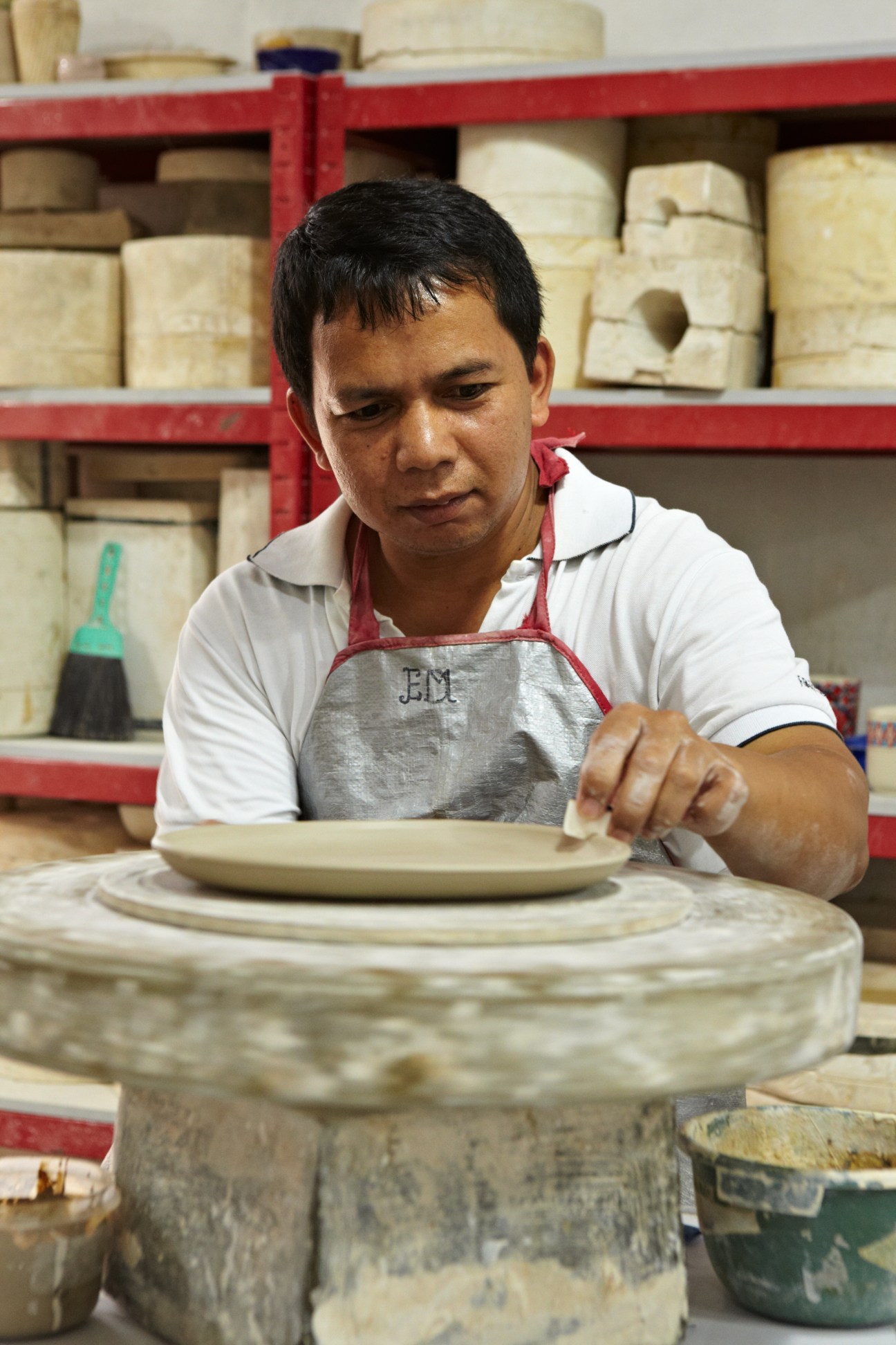 Craftsman shaping a ceramic plate at a pottery wheel in Kandura's workshop with shelves of clay vessels in the background.