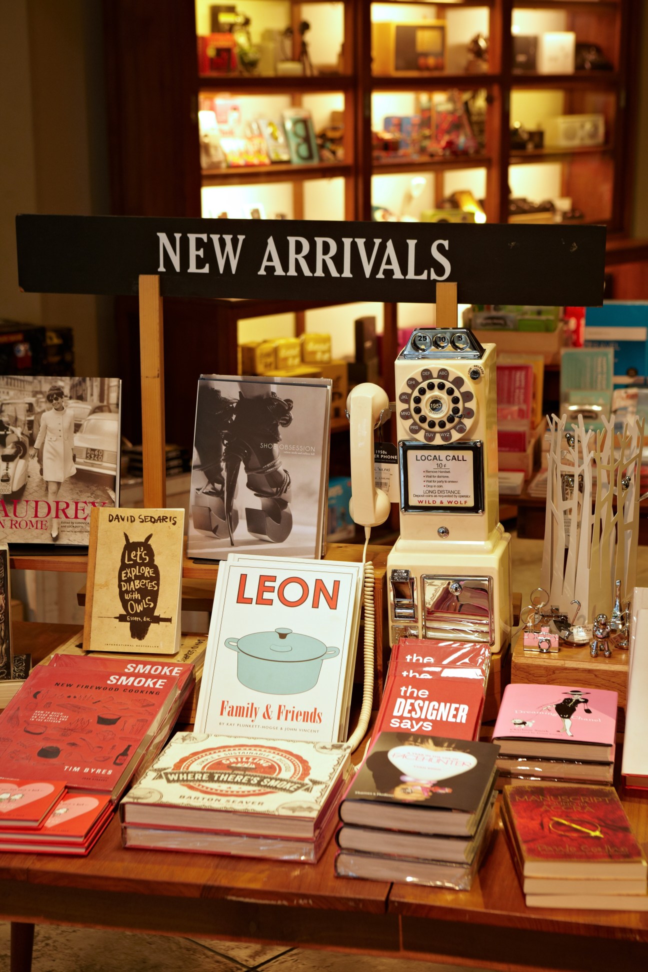 Bookshop display window showing new arrivals including design books, vintage telephone, and gift items with illuminated shelving behind.