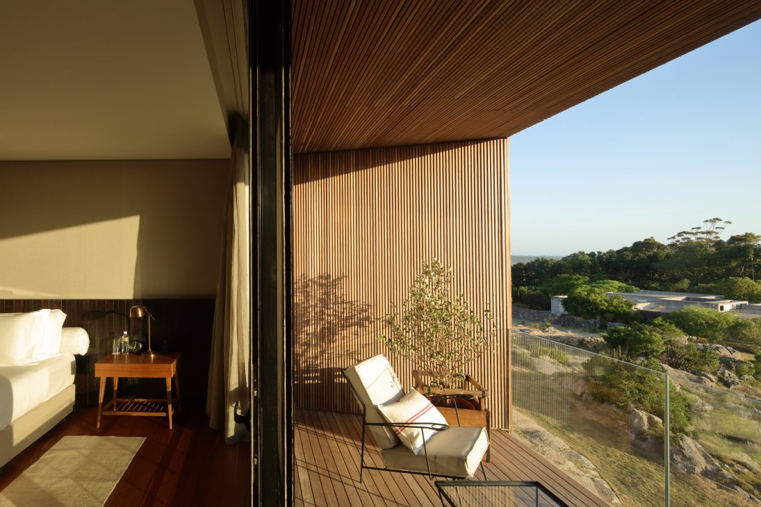 Modern hotel bedroom with wooden furniture overlooks a coastal landscape through floor-to-ceiling windows and vertical wooden cladding.