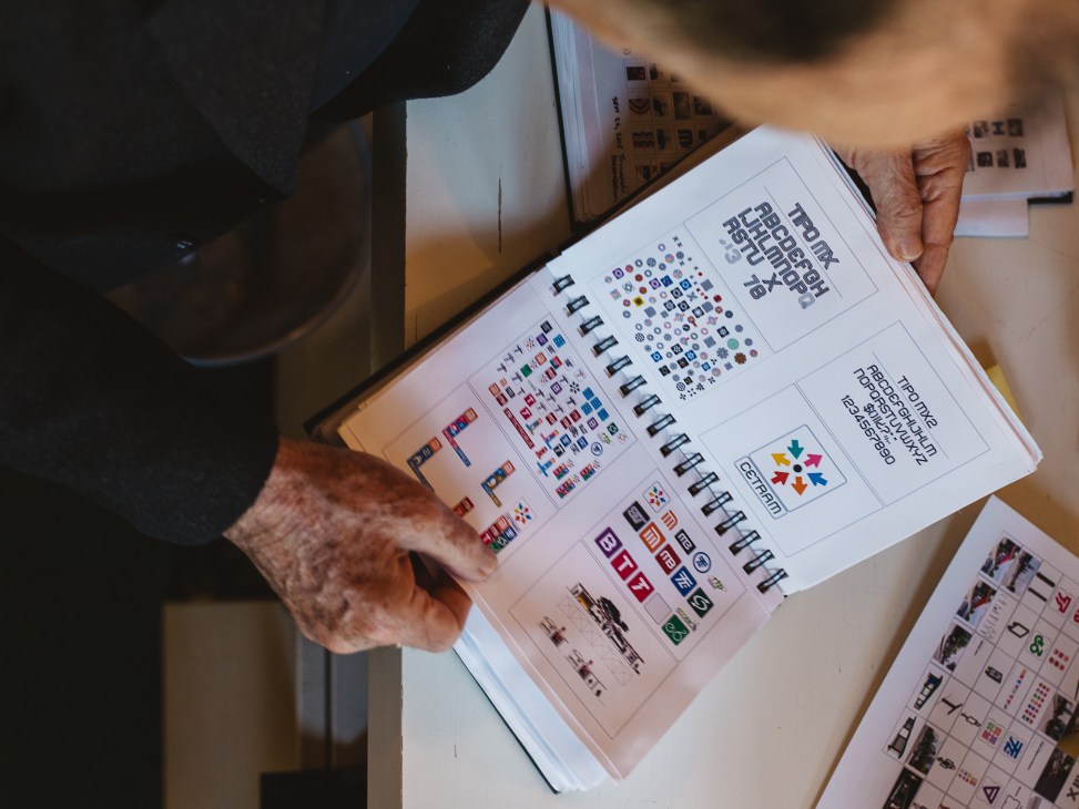 Hands holding open a sketchbook displaying various logo designs and typographic studies organized in a grid layout.