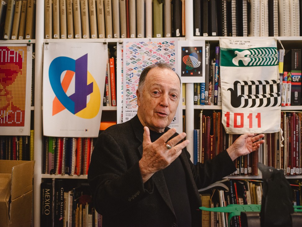 Graphic designer Lance Wyman gestures while standing in front of bookshelves displaying his colorful Olympic and design posters.