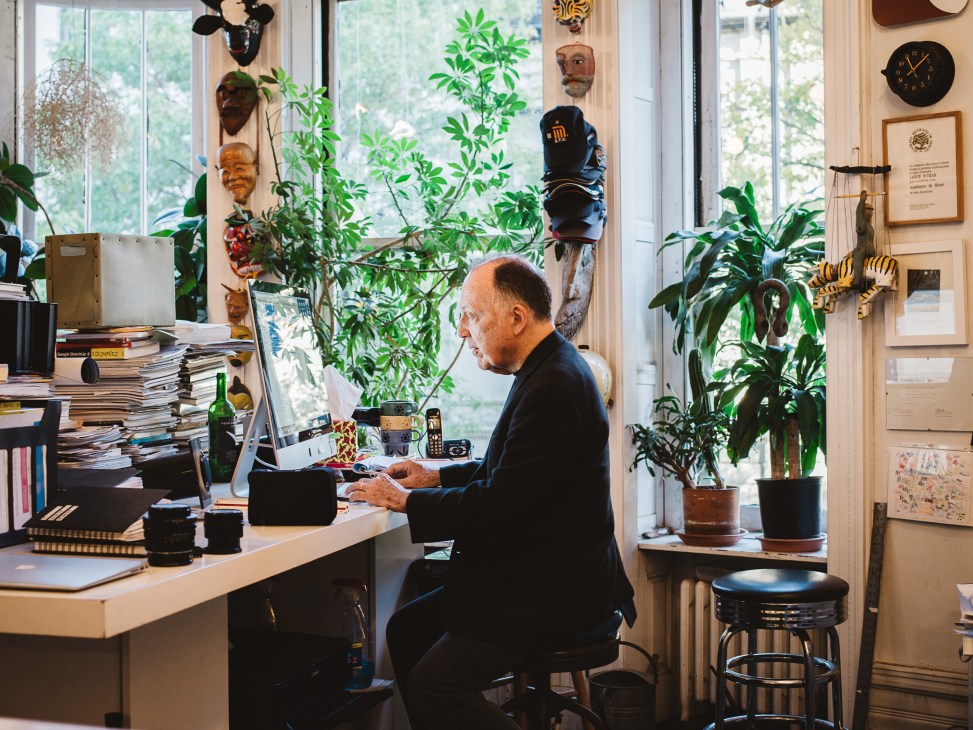 Graphic designer Lance Wyman working at his desk in his studio surrounded by plants, masks, and design materials.