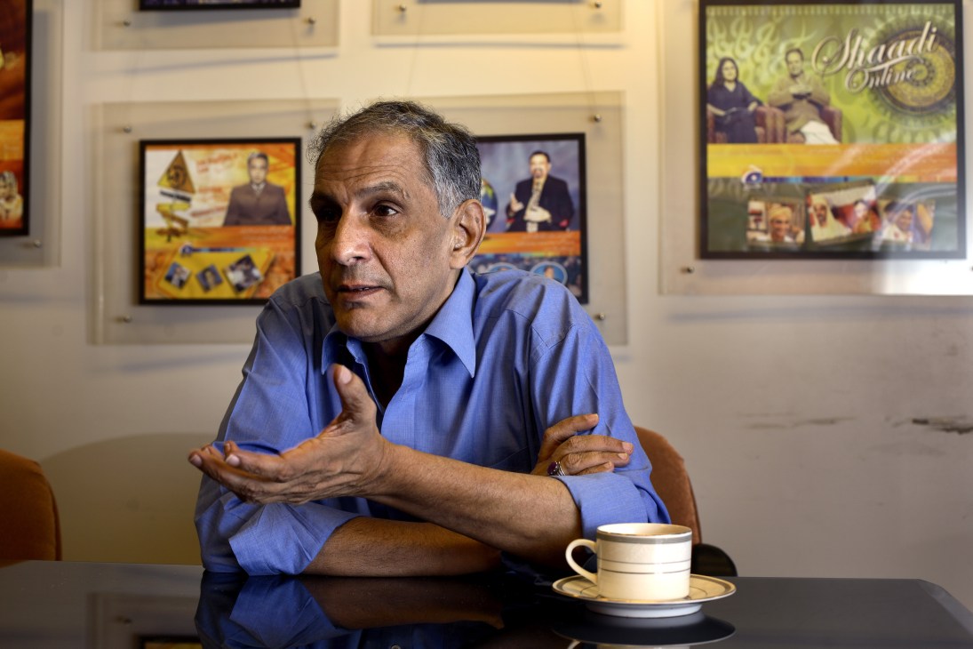 Imran Aslam, founder and president of GEO TV, gestures while speaking at a table with a cup of tea, with TV screens displaying Pakistani programming in the background.