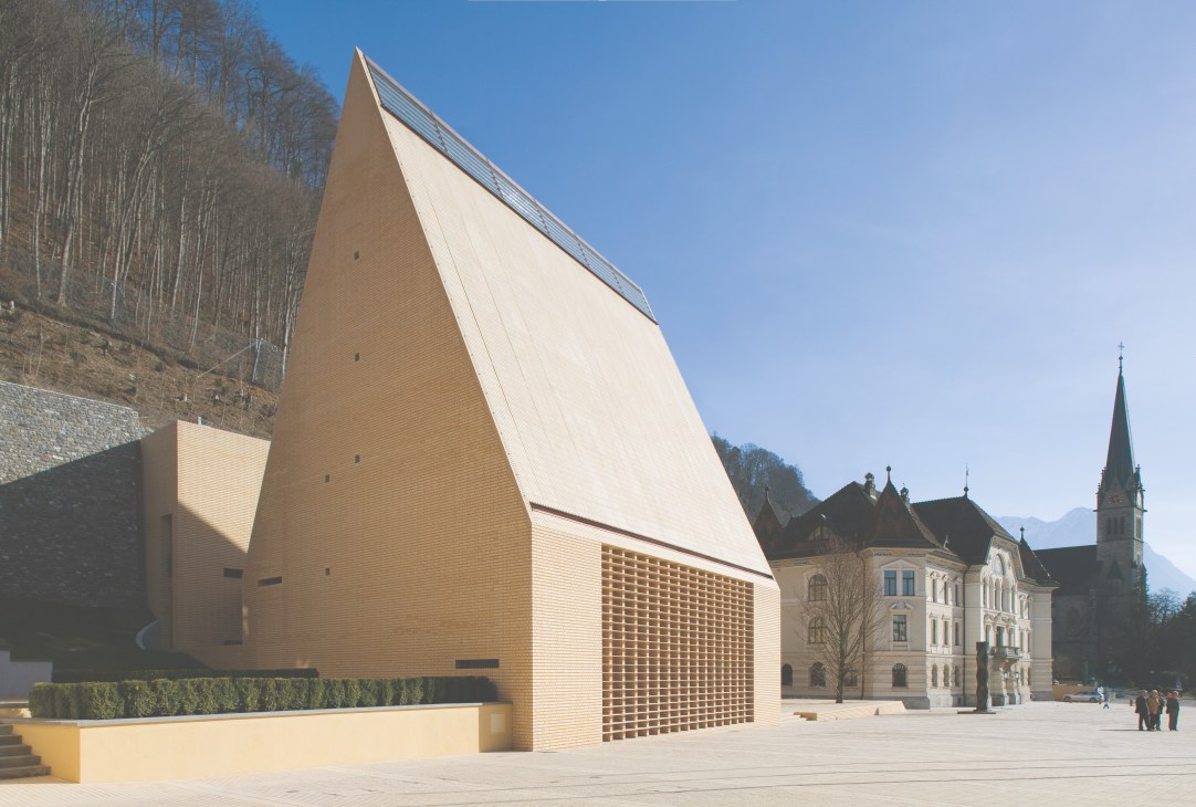 Liechtenstein's Landtag parliament building with steeply pitched roof and pale brick facade alongside a historic 1910 government building.
