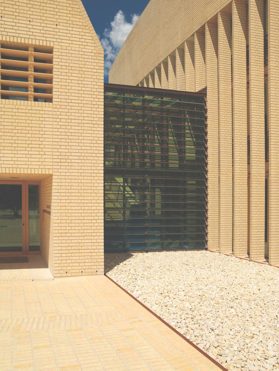 Modern brick building exterior with glass corridor connecting two structures under blue sky.