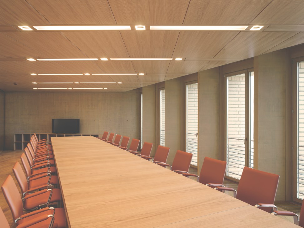Modern meeting room with long wooden table, orange chairs, wood ceiling with integrated lighting, and tall windows.