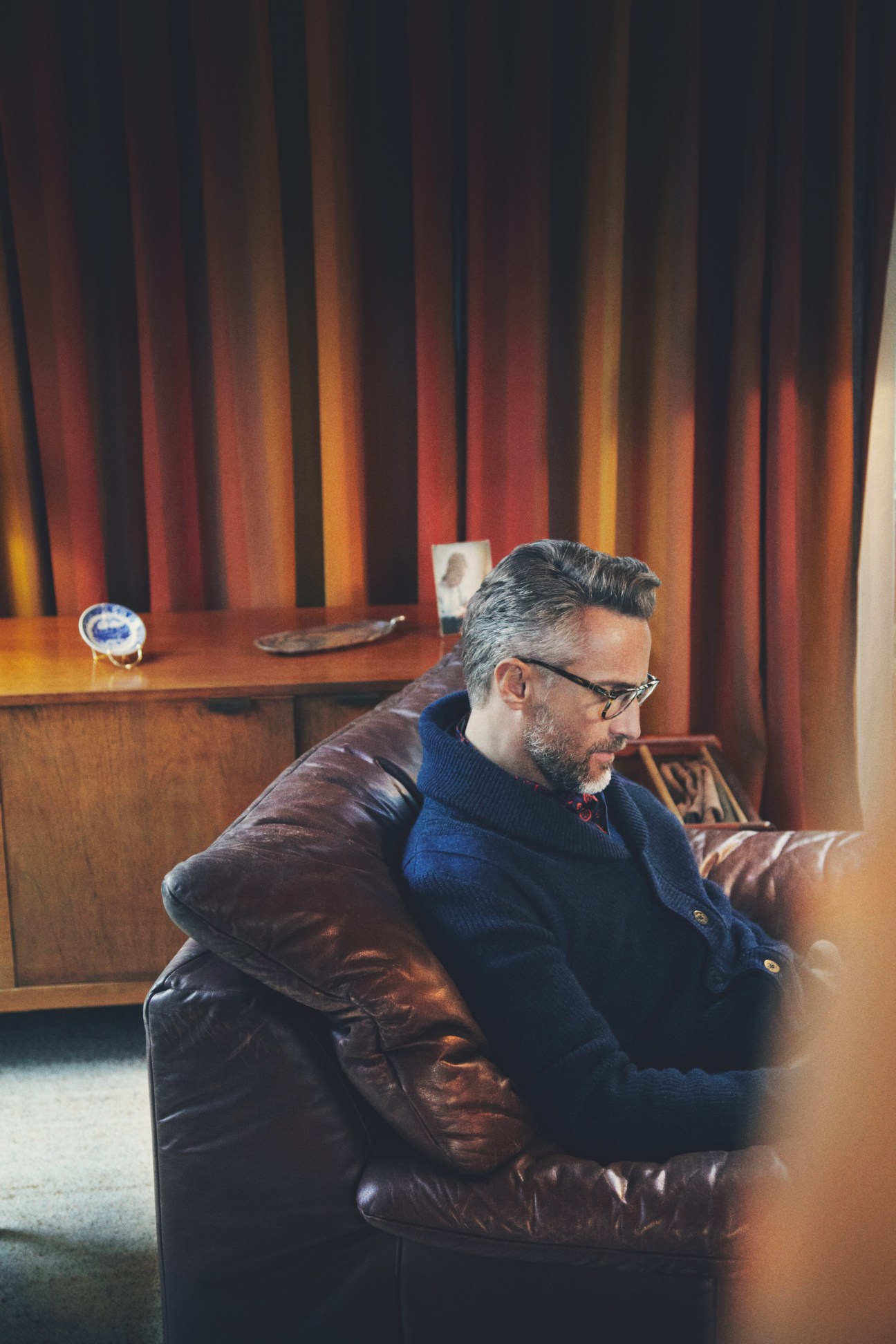 Man seated on leather sofa wearing Persol glasses, Dunhill cardigan, Bally shirt and Prada scarf in mid-century modern interior.