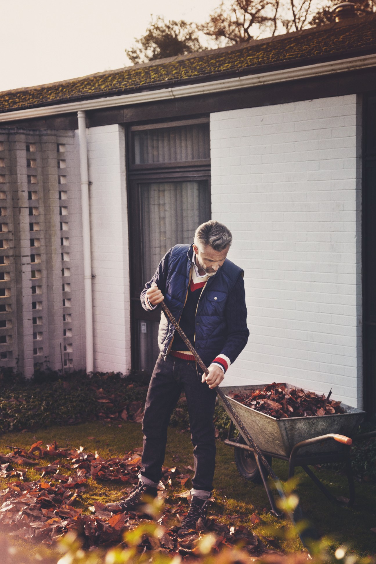 Man in navy vest and Hackett jumper raking leaves outside a modern house.