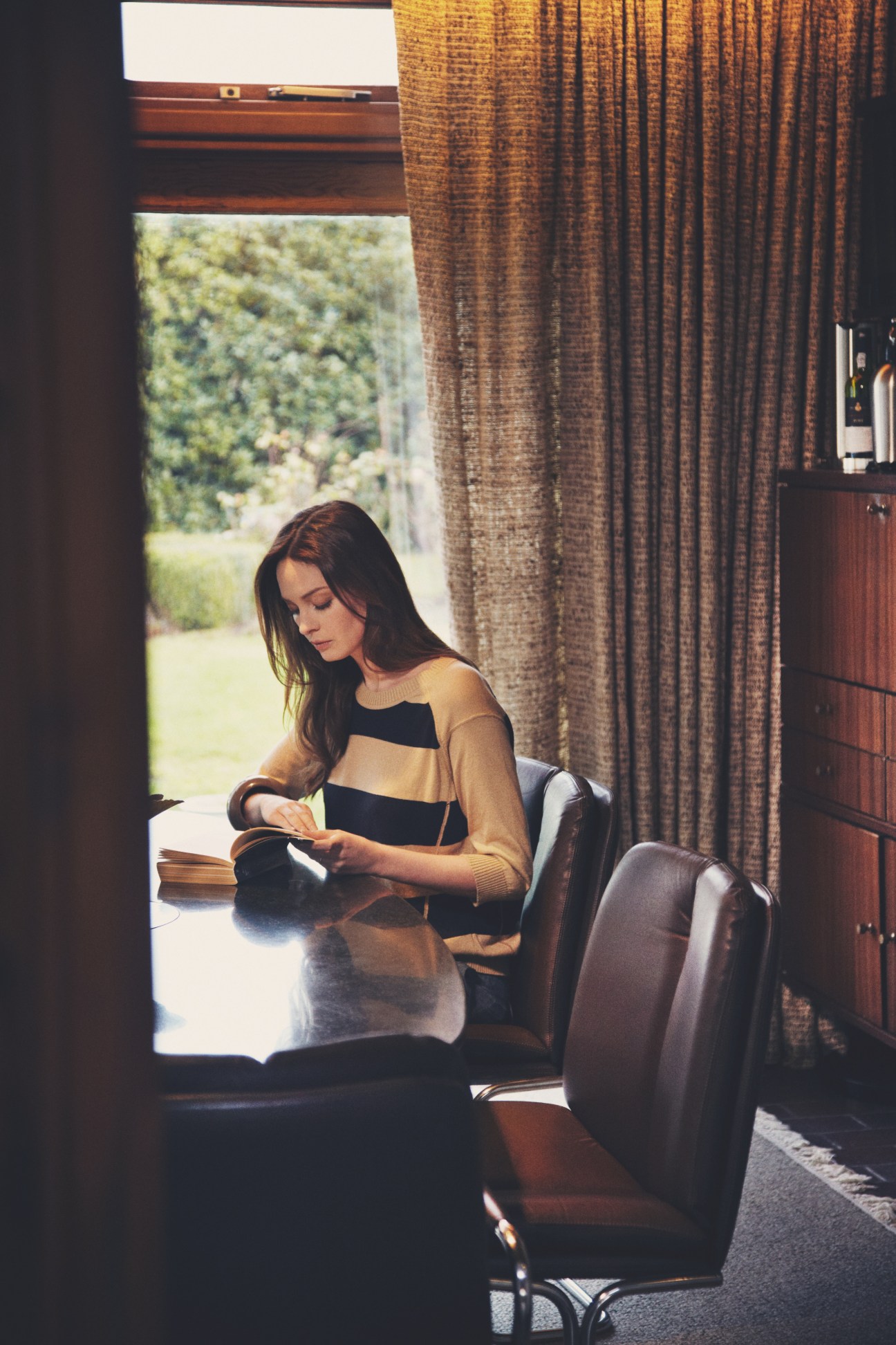 Woman in a Woolrich John Rich & Bros jumper, Hermès bracelet, and Maison Kitsuné trousers reading at a desk in a modernist interior.