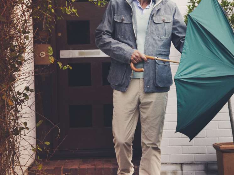 Man in denim jacket and beige trousers holding a teal umbrella outside a modern home.