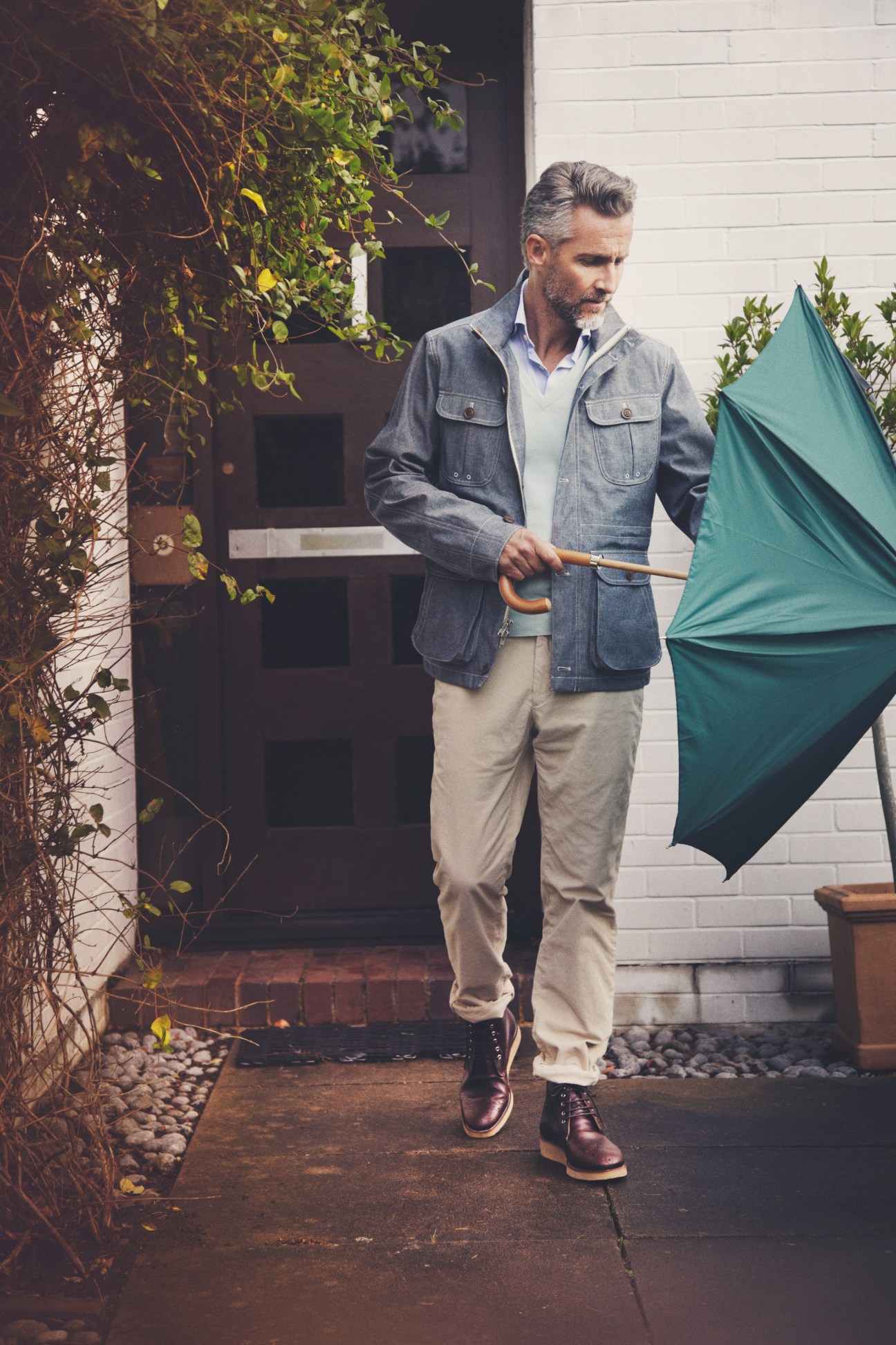Man in denim jacket and beige trousers holding a teal umbrella outside a modern home.