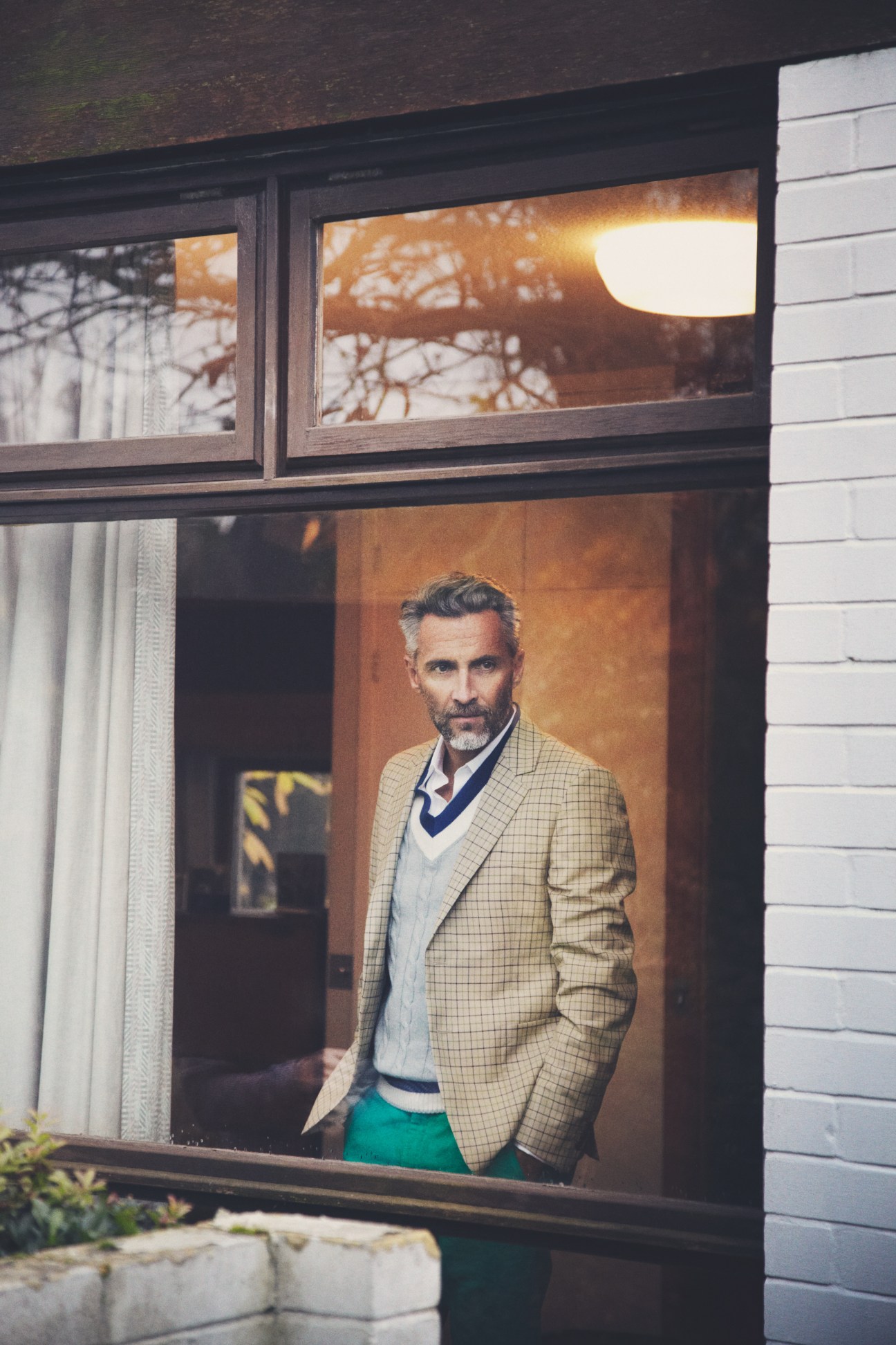 Man in cream checked Prada jacket and navy waistcoat looking out from modernist window