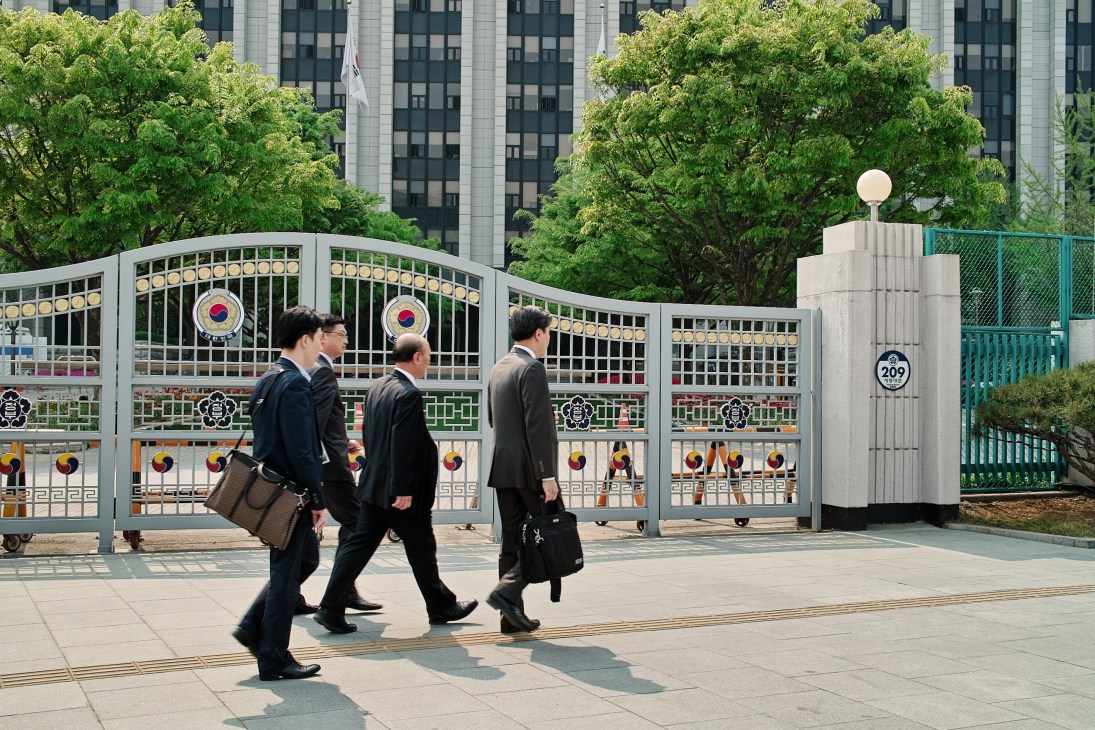 Three men in business suits walk past a secured gate with South Korean emblem at the Ministry of Unification in Seoul.