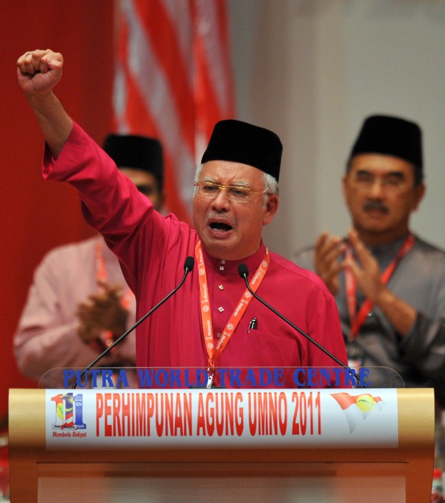 Malaysian politician in red shirt raising fist at podium during election campaign speech.